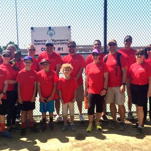 a group photograph of young girls and boys standing with a smaller group of adults, all in red shirts and standing in front of a baseball diamond